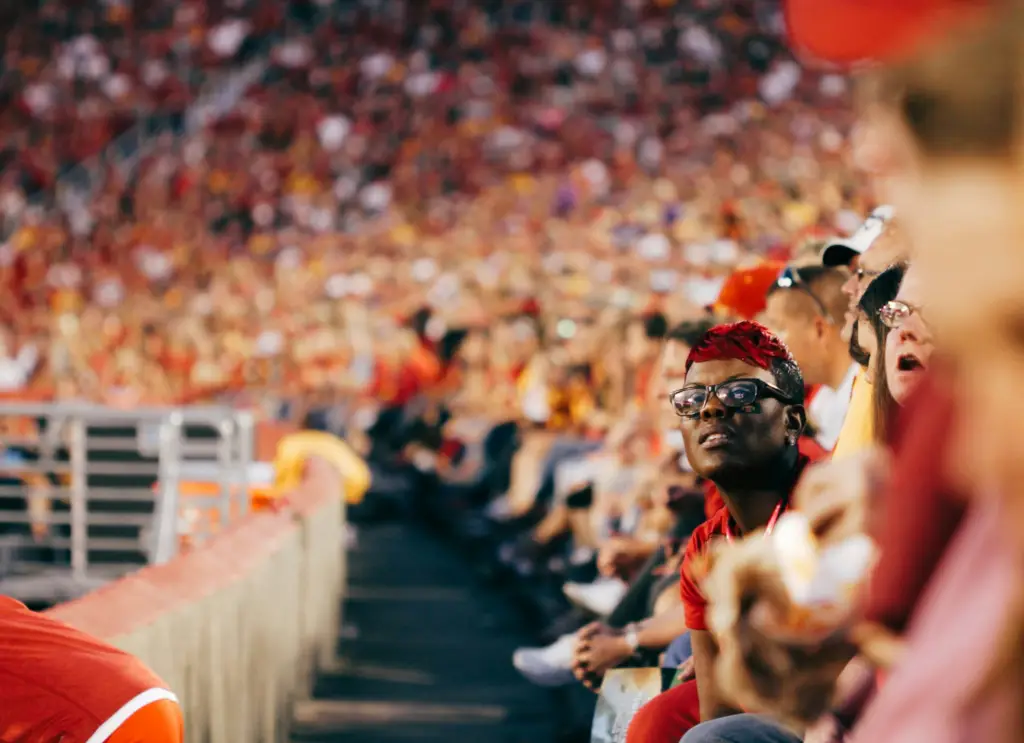 People sitting in a crowded stadium, with one fan in the foreground wearing glasses and team colors, focused on the game each used a university ticketing software platform to make their purchase.